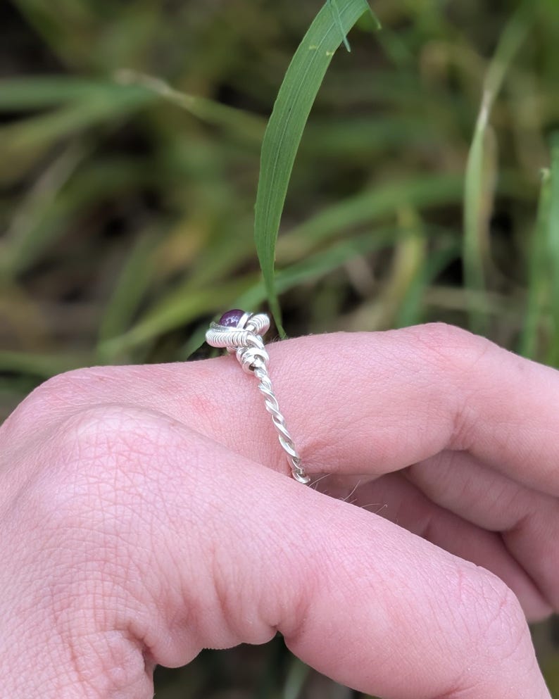 K&ouml;nnte beinhalten: Ein silberner Ring mit einem gedrehten Band und einem kleinen violetten Edelstein in der Mitte.