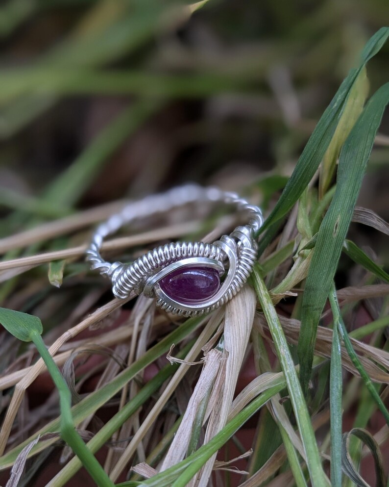 K&ouml;nnte beinhalten: Ein Ring aus silbernem Draht mit einem violetten Edelstein. Der Ring liegt auf einem Hintergrund aus gr&uuml;nem Gras.