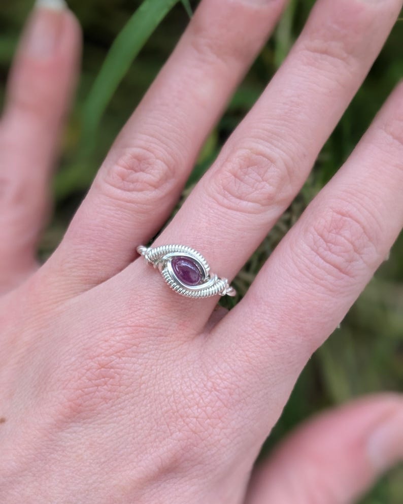 K&ouml;nnte beinhalten: Ein Ring aus silbernem Draht mit einem violetten Edelstein. Der Ring wird an einem Finger getragen.