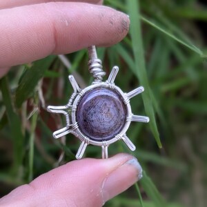 Peut inclure: Un pendentif en fil d'argent enroulé autour d'une pierre d'agate ronde, de couleur pourpre et blanche, avec un motif tourbillonnant. Le pendentif est en forme de soleil à huit pointes.