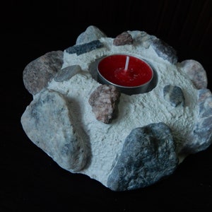 Może przedstawiać: Biały betonowy świecznik z czerwoną świeczką typu tealight w środku. Świecznik jest ozdobiony małymi szarymi i brązowymi kamieniami.