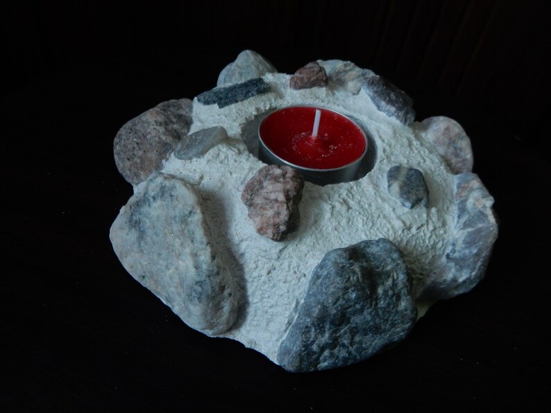 Może przedstawiać: Biały betonowy świecznik z czerwoną świeczką typu tealight w środku. Świecznik jest ozdobiony małymi szarymi i brązowymi kamieniami.