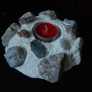 Może przedstawiać: Betonowy świecznik z czerwoną świeczką typu tealight w środku. Świecznik jest ozdobiony małymi kamieniami w r&oacute;żnych kolorach i fakturach.