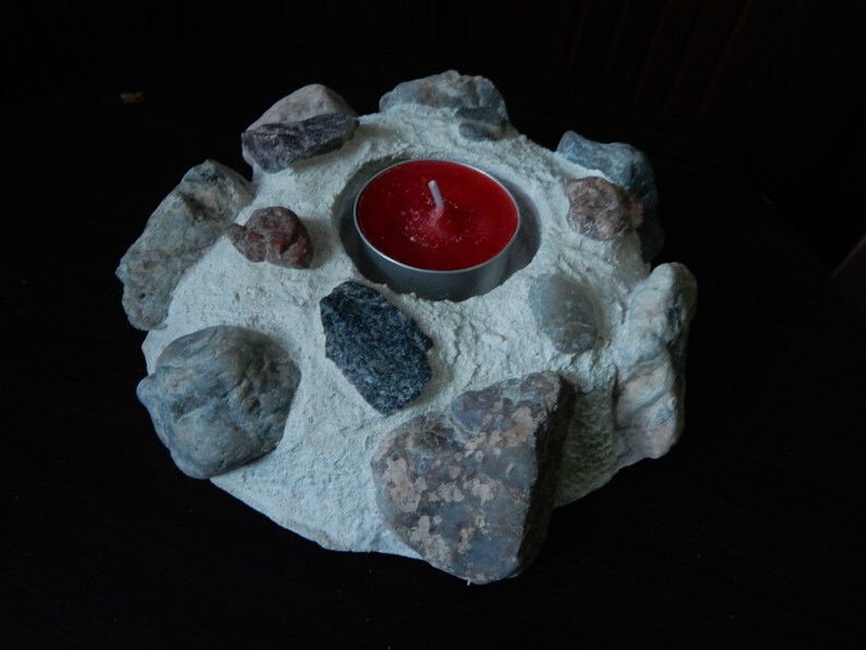Może przedstawiać: Betonowy świecznik z czerwoną świeczką typu tealight w środku. Świecznik jest ozdobiony małymi kamieniami w r&oacute;żnych kolorach i fakturach.