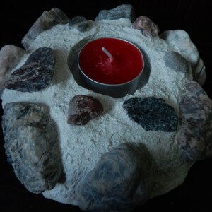 Może przedstawiać: Okrągły betonowy świecznik z czerwoną świeczką typu tealight w środku. Świecznik jest ozdobiony małymi szarymi i brązowymi kamieniami.