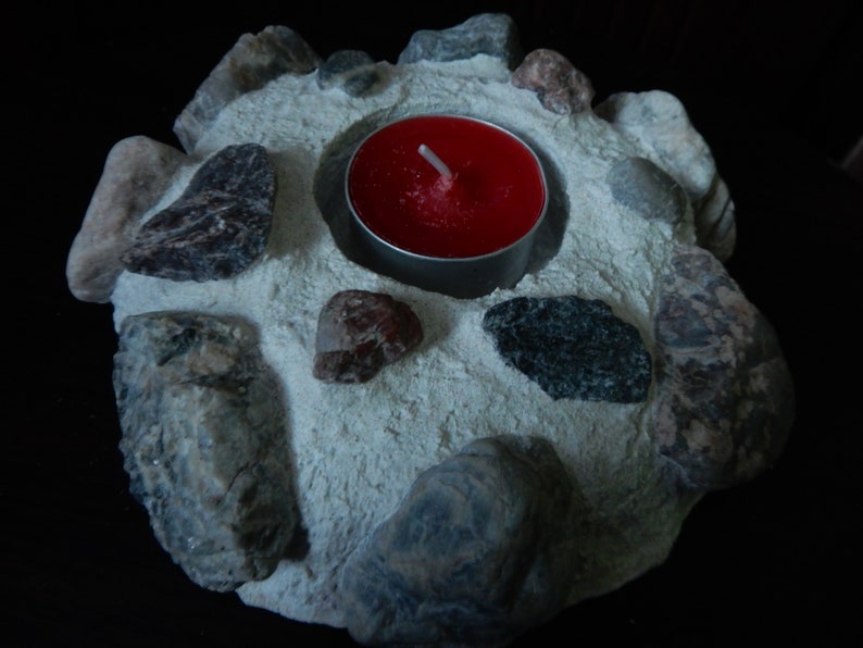 Może przedstawiać: Okrągły betonowy świecznik z czerwoną świeczką typu tealight w środku. Świecznik jest ozdobiony małymi szarymi i brązowymi kamieniami.