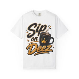 Puede incluir: Camiseta blanca con la frase "Sip on Deez" en letras negras y doradas. Se muestra una taza de café negra con un gráfico de corona dorada, con vapor saliendo del café. Prenda de vestir informal.
