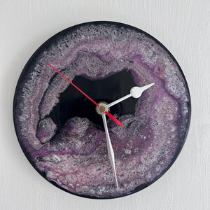 May include: A circular wall clock with a black centre surrounded by a textured, swirling ring of purple, white and black resin. The clock features two white hands and a thin red second hand attached to a central gold-toned mechanism.