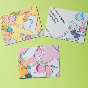 Puede incluir: Tres ilustraciones coloridas de un gato y dos conejos. El gato sostiene una taza y un lápiz. Los conejos llevan pajaritas. Las ilustraciones están en papel blanco con un fondo verde claro.