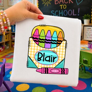May include: A white t-shirt featuring a graphic of a yellow checkered crayon box filled with five colorful crayons in pink, green, purple, blue, and orange. The box has a blue oval with the name Blair and a pink crayon labeled FOES below.