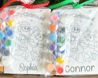 May include: Two Christmas-themed paint-your-own kits, each featuring a Santa Claus and reindeer design. The kits are packaged in clear plastic bags with red and green polka dot ribbons. The kits include paint pots, brushes, and a card with the names "Sophia" and "Connor" written on them.