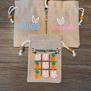 May include: Three burlap gift bags with personalized names, "Miles", "Hannah", and a third bag with the text "Happy Easter" and a tic-tac-toe game with bunny and carrot images.