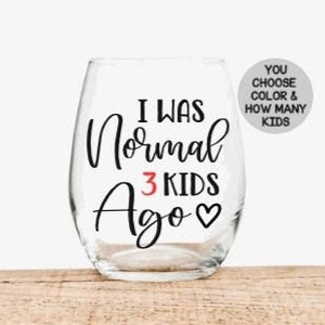 May include: A clear glass wine glass with the text "I was normal 3 kids ago" in black lettering with a red heart. The text "You choose color & how many kids" is on a white circle sticker.
