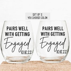 May include: Set of two clear glass wine glasses with the text "Pairs well with getting engaged 10.18.22" printed on each glass.