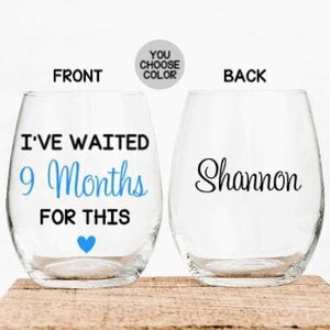 May include: A clear glass wine glass with a blue heart and the text "I've waited 9 months for this" on the front. The back of the glass has the name "Shannon" written on it.