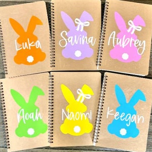 May include: Six brown spiral notebooks with bunny designs and names on each. The bunnies are in different colors: orange, purple, pink, green, yellow, and blue. The names are Luka, Savina, Aubrey, Noah, Naomi, and Keegan.