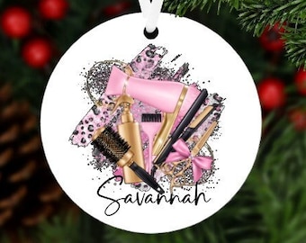 May include: A white round Christmas ornament with a pink hair dryer, gold hair straightener, gold hair brush, and gold hairspray bottle. The ornament has a glittery background and the name "Savannah" written in black cursive.