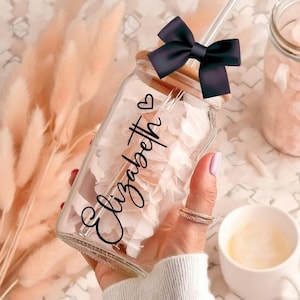 May include: A clear glass mason jar with a wooden lid and a black bow. The jar is filled with white flower petals and has the name "Elizabeth" written on it in black cursive. The jar has a straw sticking out of the lid.