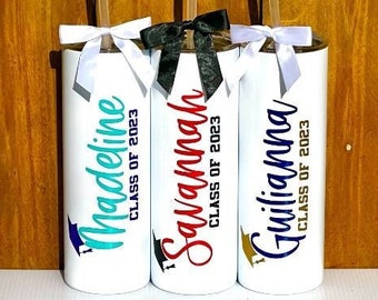 May include: Three white insulated tumblers with black lids and straws. Each tumbler has a different name and the text "Class of 2023" printed on it. The names are Madeline, Savannah, and Giuliana.