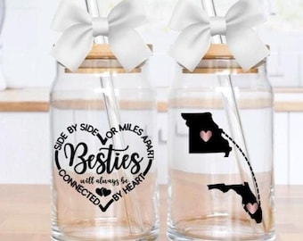 Puede incluir: Dos tarros de cristal transparente con tapas de bambú y pajitas. Los tarros tienen un diseño en blanco y negro que dice "Side by Side or Miles Apart, Besties will always be connected by heart". Un tarro tiene un contorno negro de Misuri con un corazón rosa en el medio y una línea punteada que lleva a un contorno negro de Florida con un corazón rosa en el medio.