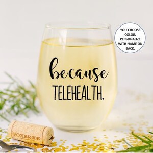 May include: A clear glass wine glass with the text "because telehealth." printed on the front. The glass is filled with a light yellow liquid. The glass is sitting on a white surface with gold glitter.