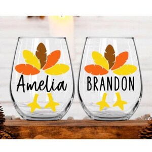 May include: Two clear glass wine glasses with a turkey design and the names "Amelia" and "Brandon" printed on them. The turkey design features orange, yellow, and brown feathers.