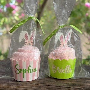 May include: Two pink cupcake-shaped gifts, each wrapped in clear cellophane and tied with a green polka dot ribbon. Each cupcake has a bunny ear topper and a personalized name on the cupcake liner. One liner is pink and white striped with the name "Sophia", the other is green with the name "Brielle".