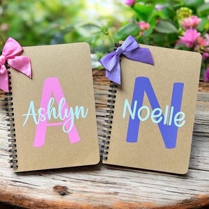 May include: Two brown spiral notebooks with personalized names. The notebook on the left has a pink bow and the name "Ashlyn" with a pink letter A. The notebook on the right has a purple bow and the name "Noelle" with a purple letter N.