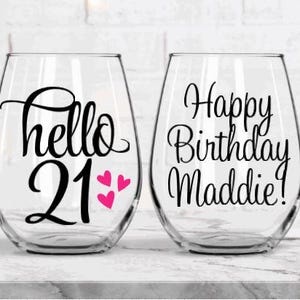 May include: Two clear stemless wine glasses. One glass has the text "hello 21" with three pink hearts. The other glass has the text "Happy Birthday Maddie!". Both glasses are for a birthday celebration.
