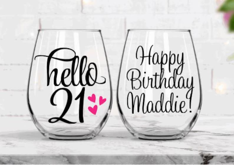 May include: Two clear stemless wine glasses. One glass has the text "hello 21" with three pink hearts. The other glass has the text "Happy Birthday Maddie!". Both glasses are for a birthday celebration.