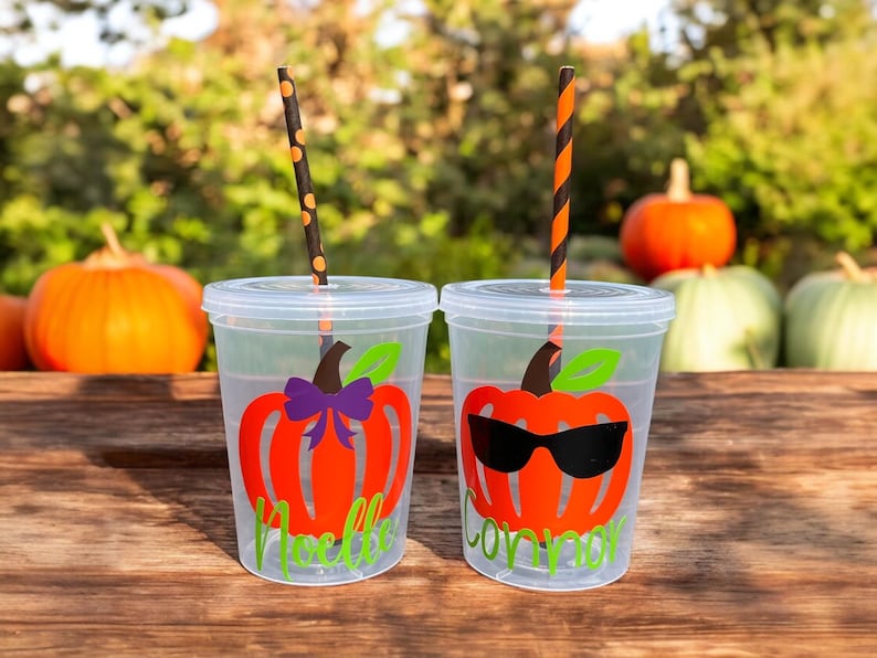 May include: Two clear plastic cups with orange pumpkin designs and black and white striped straws. One cup has a purple bow on the pumpkin and the text "Noelle" below. The other cup has sunglasses on the pumpkin and the text "Connor" below.