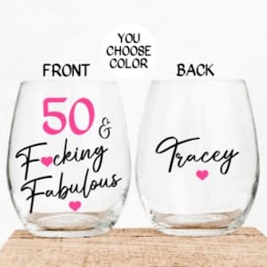 May include: Two clear wine glasses with a pink and black design. The front glass says "50 & F*cking Fabulous" with pink hearts. The back glass says "Tracey" with a pink heart.