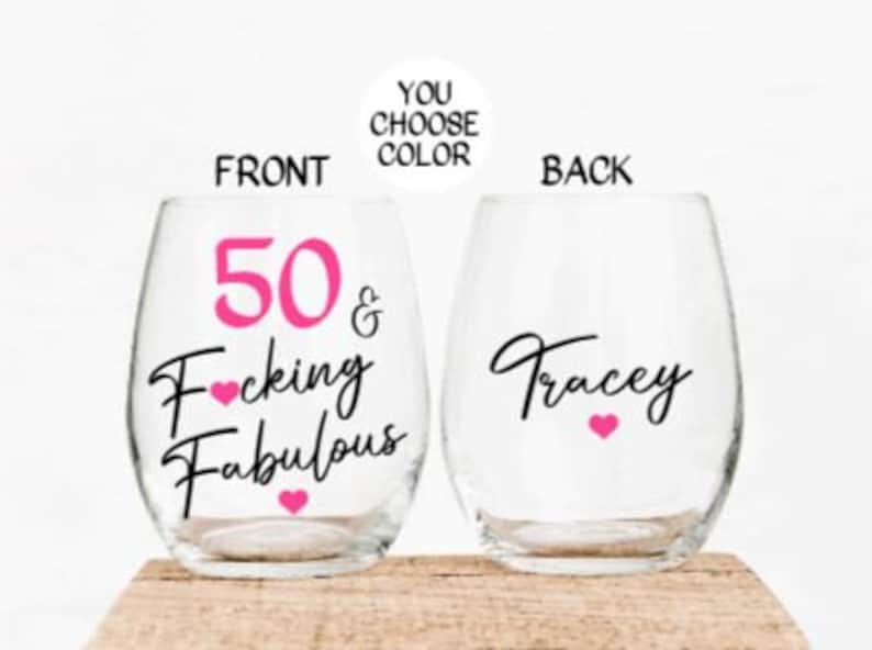 May include: Two clear wine glasses with a pink and black design. The front glass says "50 & F*cking Fabulous" with pink hearts. The back glass says "Tracey" with a pink heart.