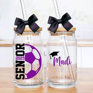 May include: Two glass jars with bamboo lids and black bows. The jars have straws and are decorated with graduation caps, a soccer ball, and the text "Senior 2025" and "Madi".