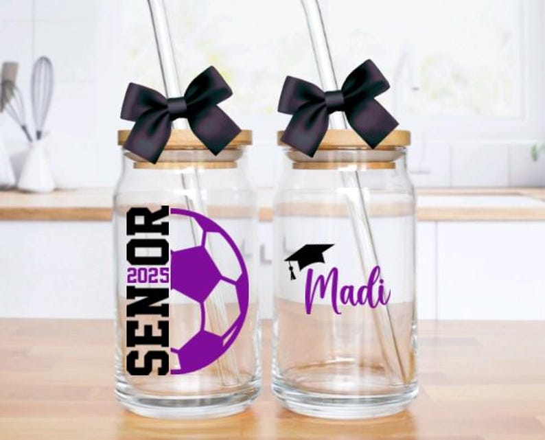 May include: Two glass jars with bamboo lids and black bows. The jars have straws and are decorated with graduation caps, a soccer ball, and the text "Senior 2025" and "Madi".