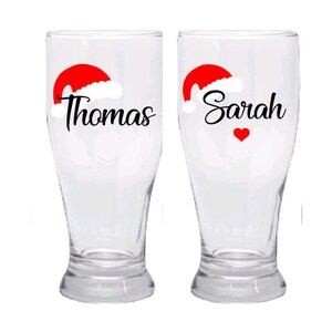 May include: Two clear glass beer mugs with red Santa hats and the names "Thomas" and "Sarah" printed on them. A red heart is printed on the mug with the name "Sarah".