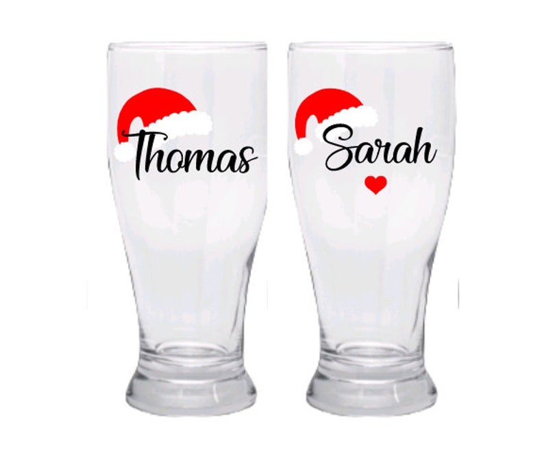 May include: Two clear glass beer mugs with red Santa hats and the names "Thomas" and "Sarah" printed on them. A red heart is printed on the mug with the name "Sarah".
