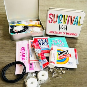 May include: A small, open silver tin containing various survival kit items, including a toothbrush, bandages, and hair ties. A second tin with the words "SURVIVAL KIT" and "OPEN IN CASE OF EMERGENCY" is next to it. Other items include lip balm and safety pins.