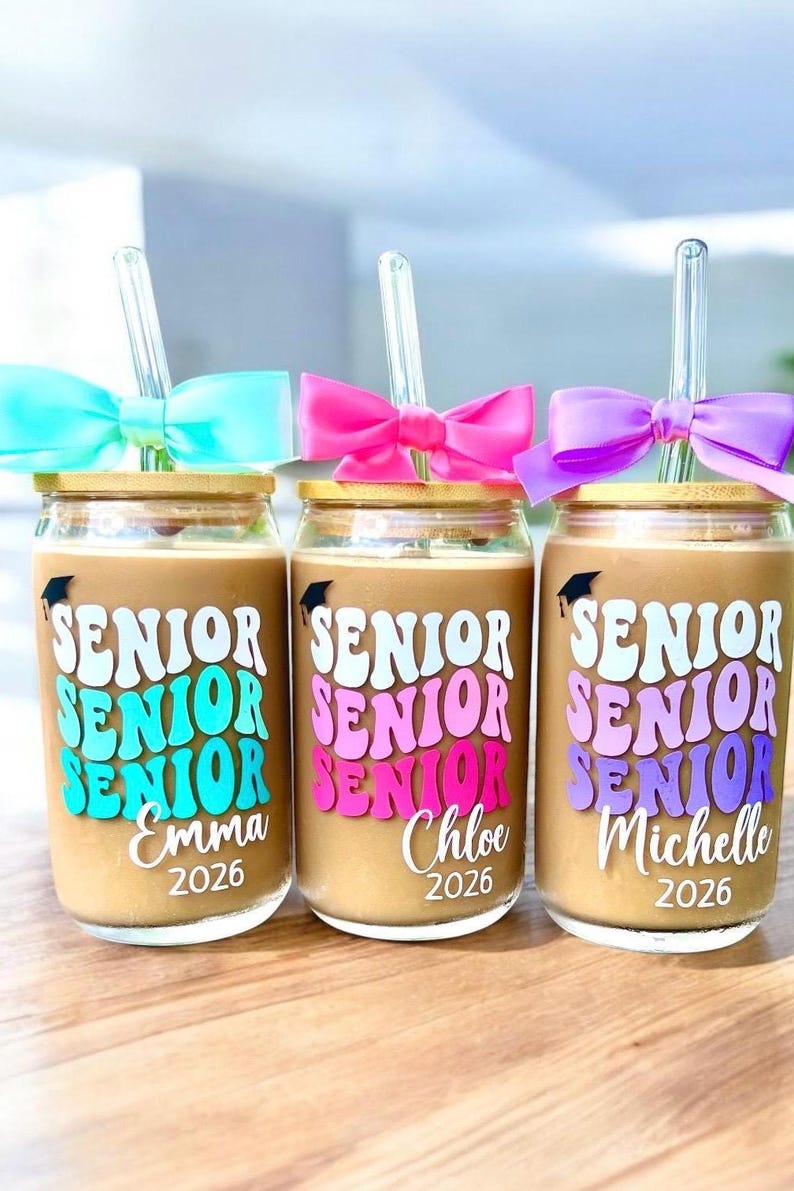 May include: Three glass tumblers with bamboo lids and clear glass straws, each featuring a graduation cap icon, the word SENIOR in wavy lettering, a personalized name, and the year 2026. Each glass is adorned with a colorful satin bow.