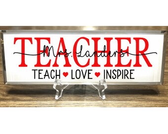 May include: A white rectangular sign with a silver frame and a black stand. The sign says "TEACHER" in red letters with "Mrs. Landers" written in cursive underneath. The bottom of the sign says "TEACH ♥ LOVE ♥ INSPIRE" in black letters.