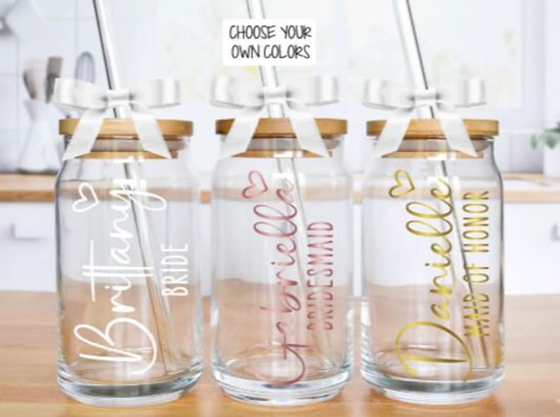 May include: Three glass mason jars with bamboo lids and white ribbon bows. Each jar has a straw and a personalised name with a title: "Brittany Bride", "Danielle Bridesmaid", and "Danielle Maid of Honour".