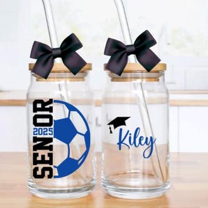 May include: Two glass mason jars with black bows and straws. One jar has a soccer ball and the text "Senior 2025". The other jar has a graduation cap and the name "Kiley".