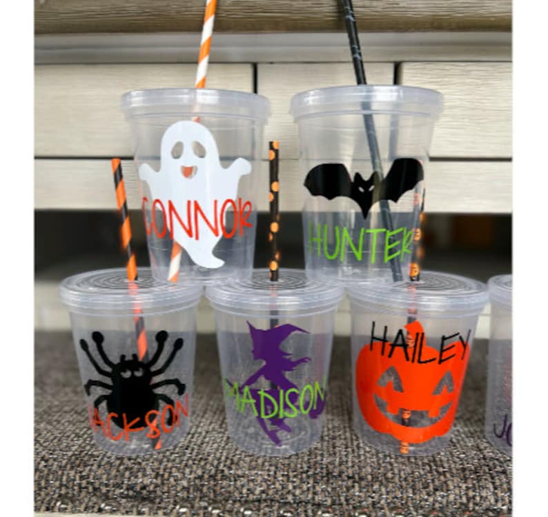 May include: Five clear plastic cups with personalized Halloween designs and names. The designs include a ghost, a bat, a spider, a witch, and a pumpkin. Each cup has a black and orange striped straw.