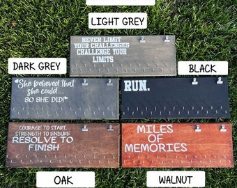 May include: Wooden medal display boards in various stain colours: light grey, dark grey, black, oak, and walnut. Each board has motivational text in white and small clips for hanging medals. The boards are displayed on grass.