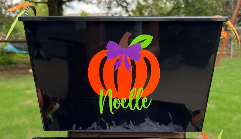 May include: Black plastic storage bin with a pumpkin design and the name "Noelle" in green lettering. The pumpkin is orange with a green stem and a purple bow.