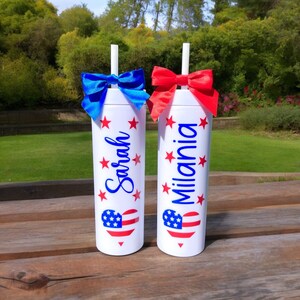 May include: Two white tumblers with red, white, and blue American flag hearts and stars. The tumblers have the names "Sarah" and "Milania" written on them. Each tumbler has a blue or red bow and a straw.