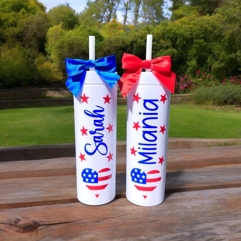 May include: Two white tumblers with red, white, and blue American flag hearts and stars. The tumblers have the names "Sarah" and "Milania" written on them. Each tumbler has a blue or red bow and a straw.