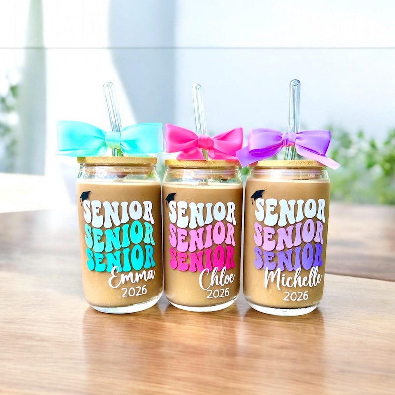 May include: Three glass tumblers with bamboo lids and clear glass straws, each topped with a colorful bow. The glasses feature the word SENIOR in a wavy font, a small graduation cap icon, a personalized name, and the year 2026 in white text.