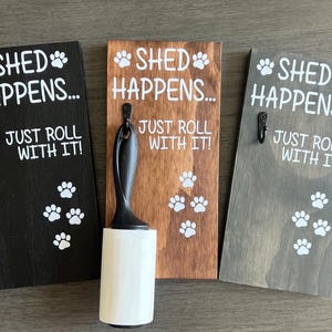 May include: Three rectangular wooden wall plaques in black, brown, and gray, each featuring a black metal hook and white text reading SHED HAPPENS... JUST ROLL WITH IT! with paw print graphics. A white adhesive lint roller hangs from the center plaque.