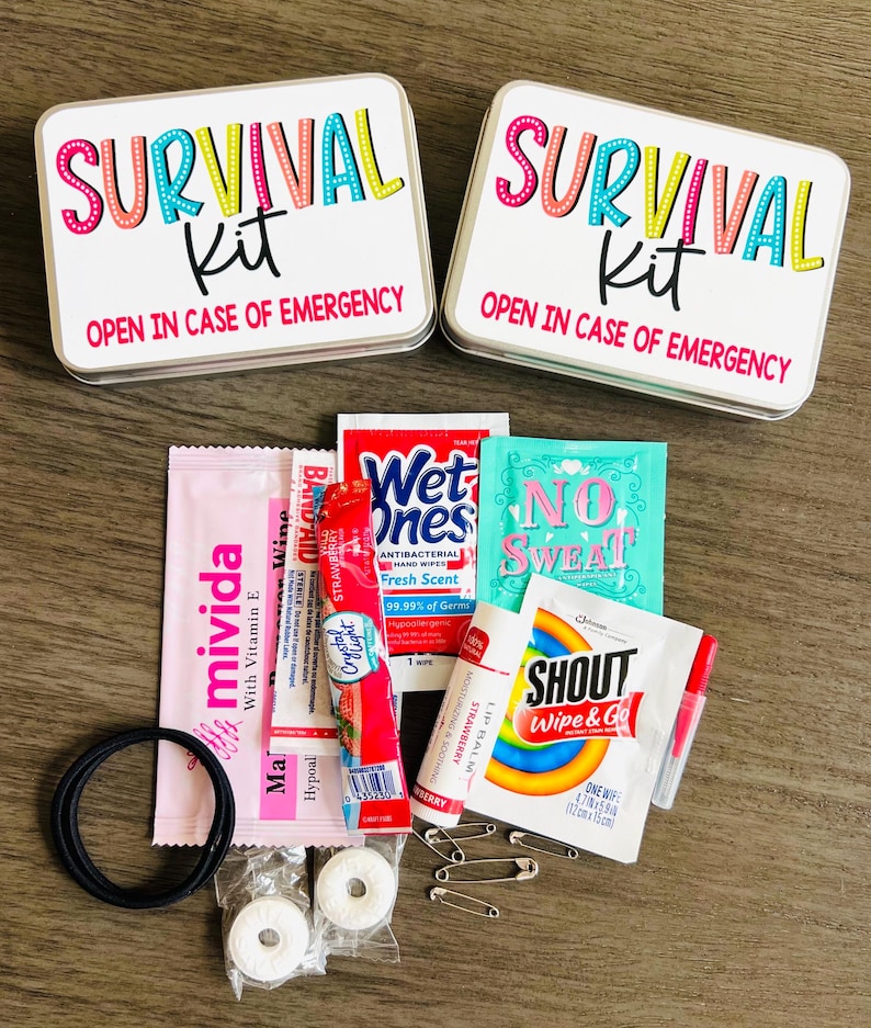 May include: Two white rectangular tins with the words "SURVIVAL Kit" in colourful text and the phrase "OPEN IN CASE OF EMERGENCY." Various small packets and items are arranged around the tins, including a hair tie, safety pins, and hygiene products.